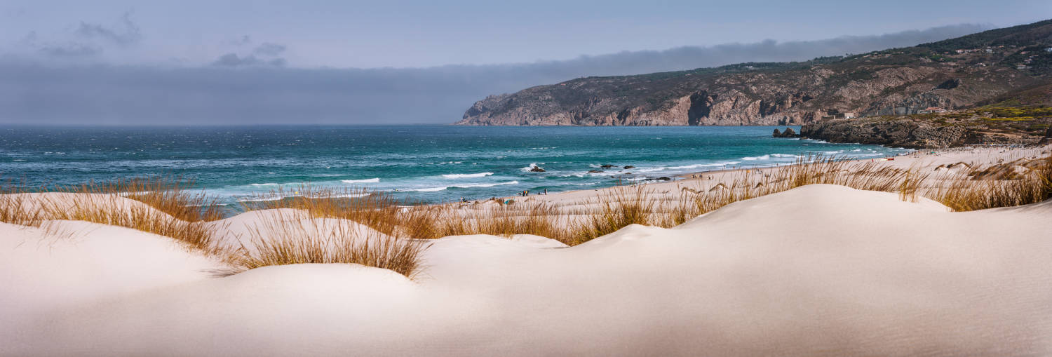 Vista panorámica de la Praia do Guincho, en Cascais, Portugal. Dunas en primer plano, océano y sierra al fondo.