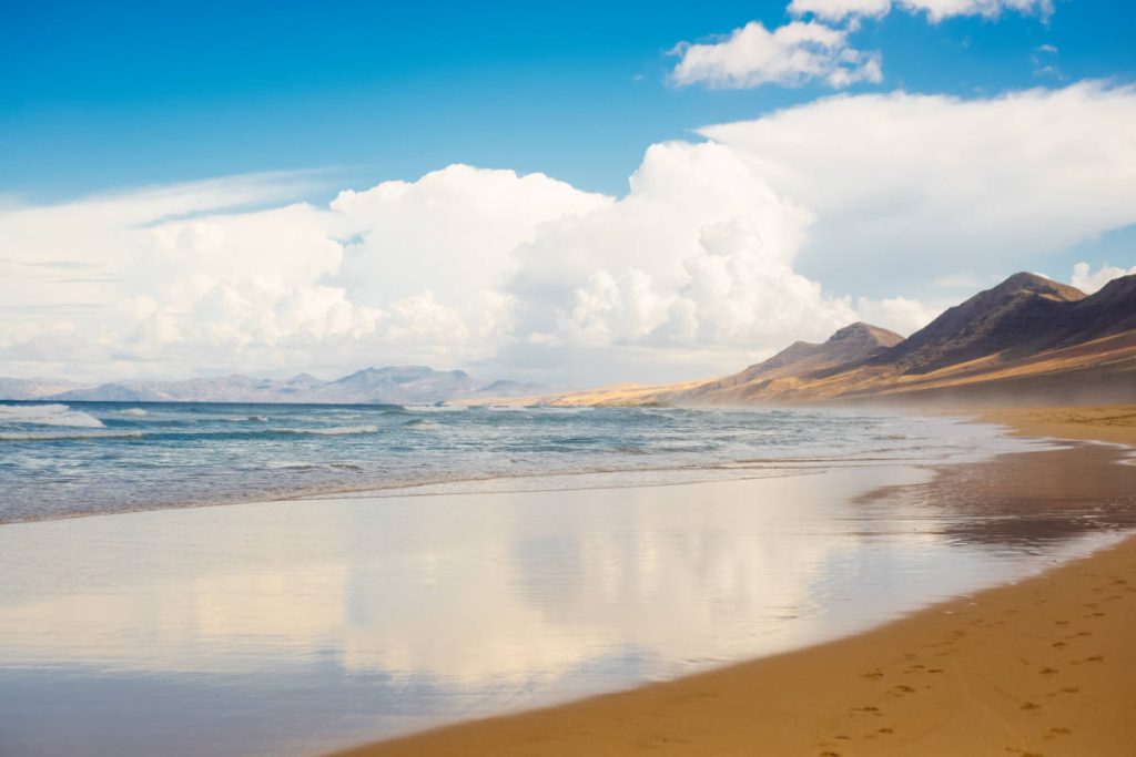 Vista de la playa de El Cofete hacia el este, con la arena mojada en primer plano y las montañas al fondo.