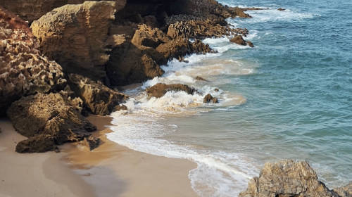 Playa de Almograve, en el Alentejo. Aparece algo de arena en el primer plano, rocas a la izquierda de la imagen, y el mar rompiendo en las rocas ocupa el resto de la foto.