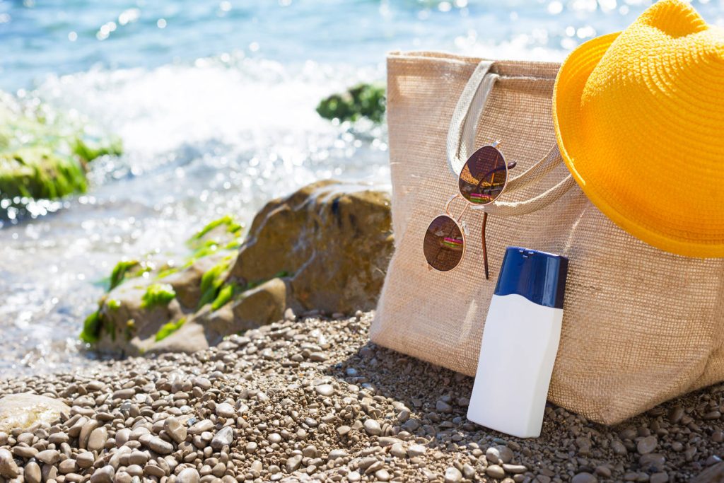 Elementos de protección solar colocados sobre la arena en la playa. Aparecen un bolso de playa, unas gafas de sol, un sombrero y un protector solar.