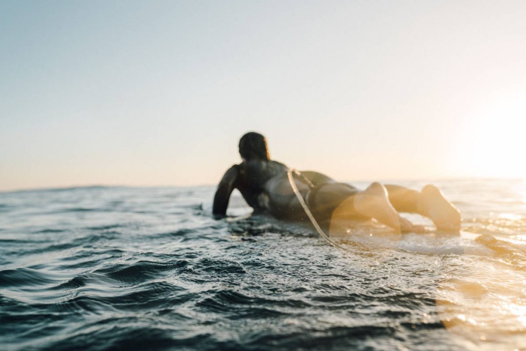 Mujer tumbada sobre una tabla de surf nadando mar adentro al atardecer.