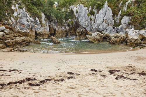 Vista de la playa interior de Gulpiyuri, en Llanes. Aparece la arena en primer plano, el agua con la marea baja, y las rocas que cierran la playa al fondo.