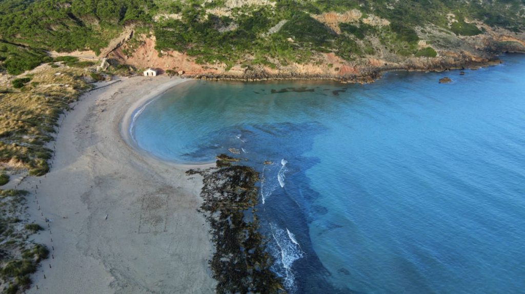 Vista aérea de la playa de Algaiarens, en Menorca. Aparece el mar de color azul turquesa a la derecha, la arena a la izquierda, y al fondo una casita y el monte cubierto de pinos.