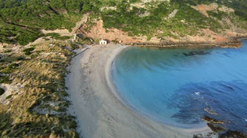 Vista aérea de la playa de Algaiarens, en Menorca. Aparece el mar de color azul turquesa a la derecha, la arena y el monte con hierba seca a la izquierda, y al fondo una casita y el monte cubierto de pinos.