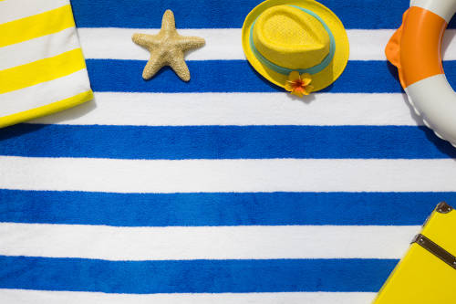 Objetos de playa en tonos amarillos sobre una toalla de rayas azules y blancas. Se aprecia una estrella de mar, un sombrero, y una esquina de un bolso, un flotador y una maleta.