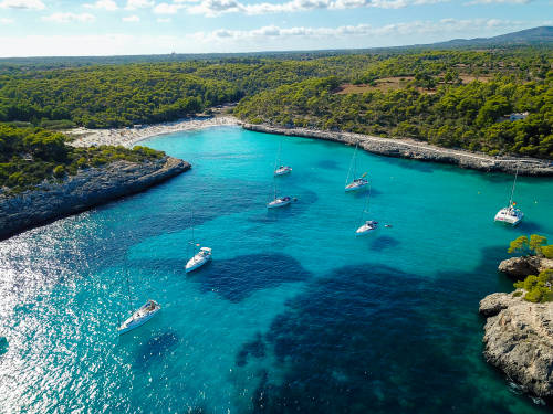 Vista aérea de Cala Llombards, con los barcos flotando en el agua y los pinares al fondo.