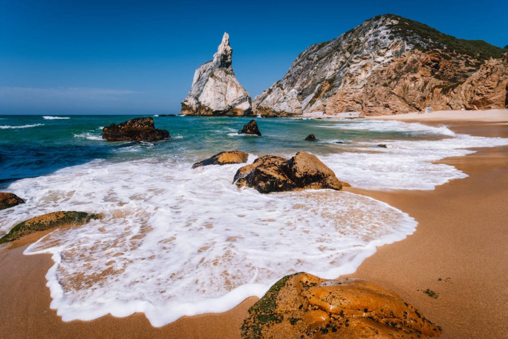 Vista de la Praia da Ursa. Arena dorada salpicada de rocas y cubierta en parte por la espuma de una ola. Al fondo, el océano turquesa y las formaciones rocosas de la costa.