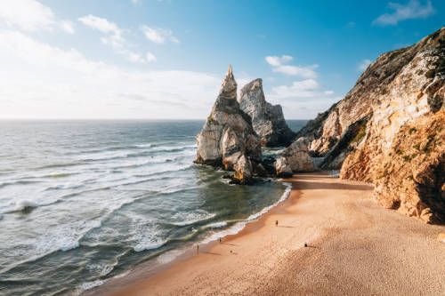 Vista aérea de la Praia da Ursa, con el agua y la arena en primer plano y el acantilado y las formaciones rocosas de la "osa y el osezno" al fondo.