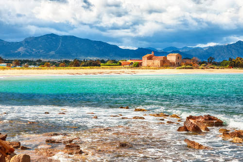 Vista de la playa de Nora, con la iglesia medieval de San Efisio junto a la orilla y las montañas al fondo