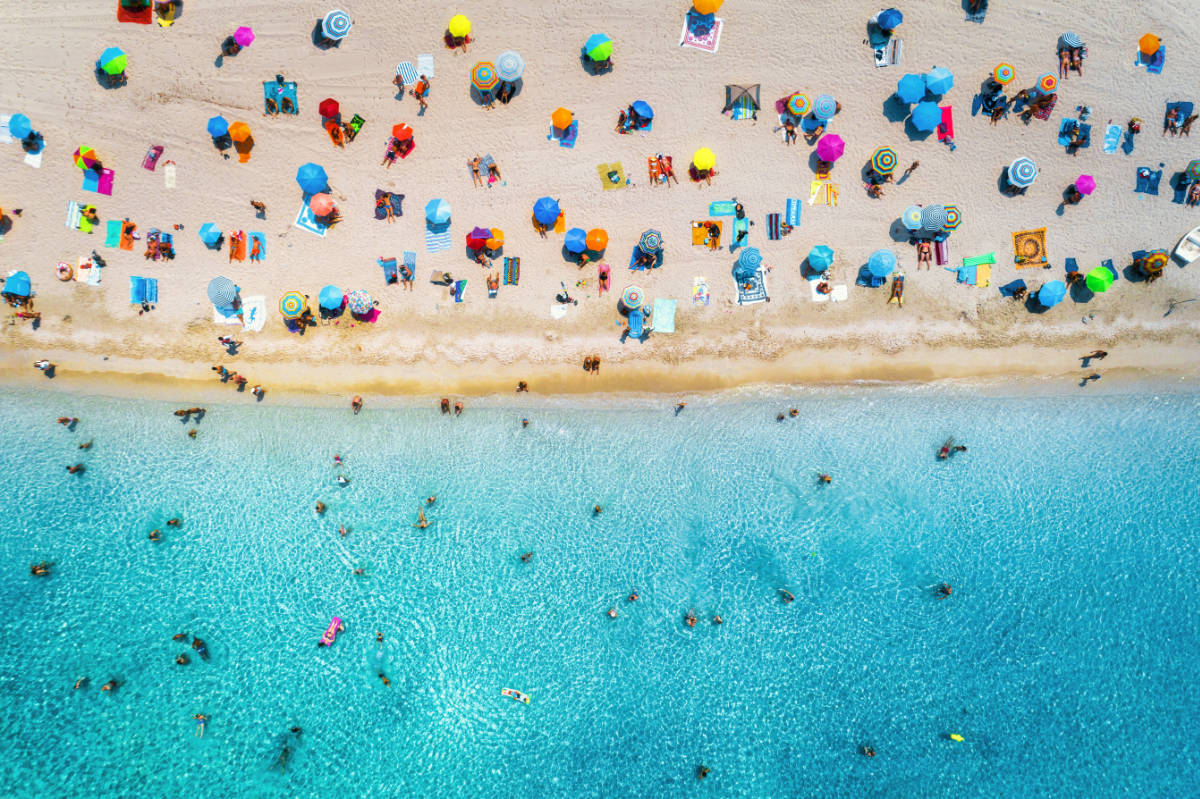 Vista aérea de una playa de arena con muchas sombrillas de colores y el mar de color turquesa con gente bañándose.